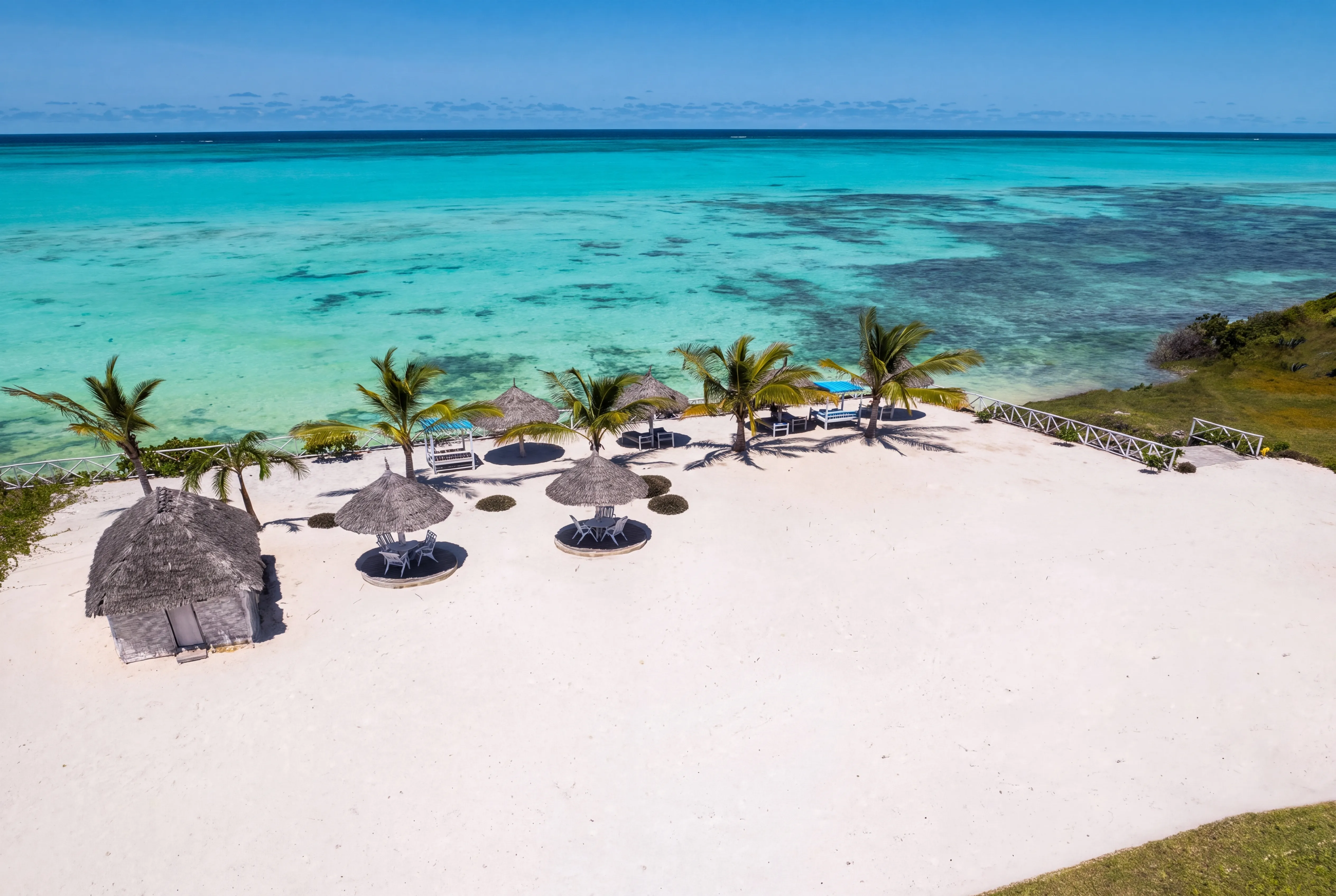 Aerial view of the Jambiani shoreline at low tide: thatched palapas along the sand, the pale green lagoon, and the white line of the reef breaking on the horizon.